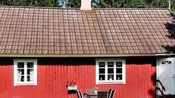 A red wooden house with a tiled roof, two windows, a door, and a small patio with a table and chairs.