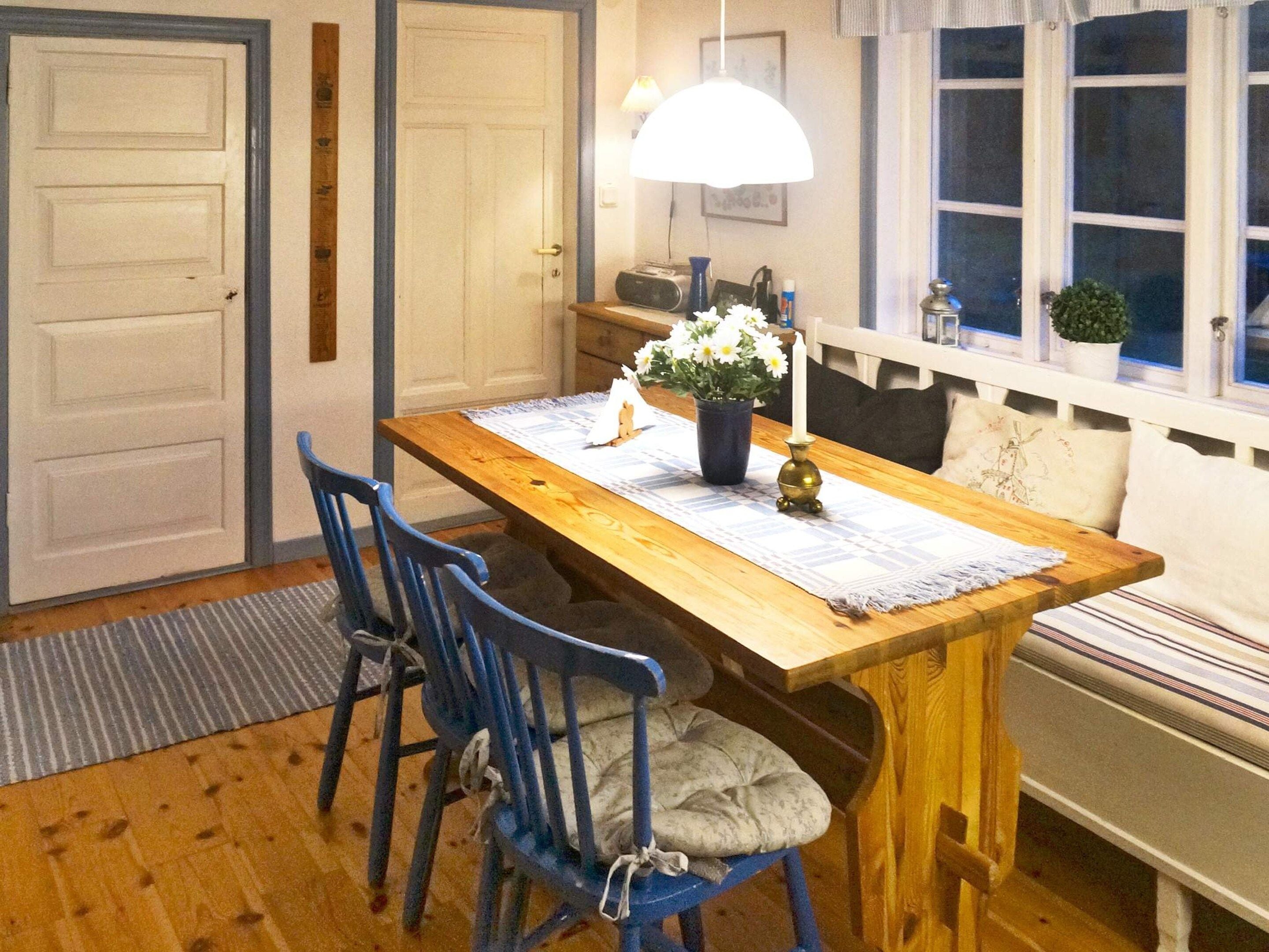 A dining area with a wooden table, blue chairs, a vase with flowers, a candle, and a window with a view.