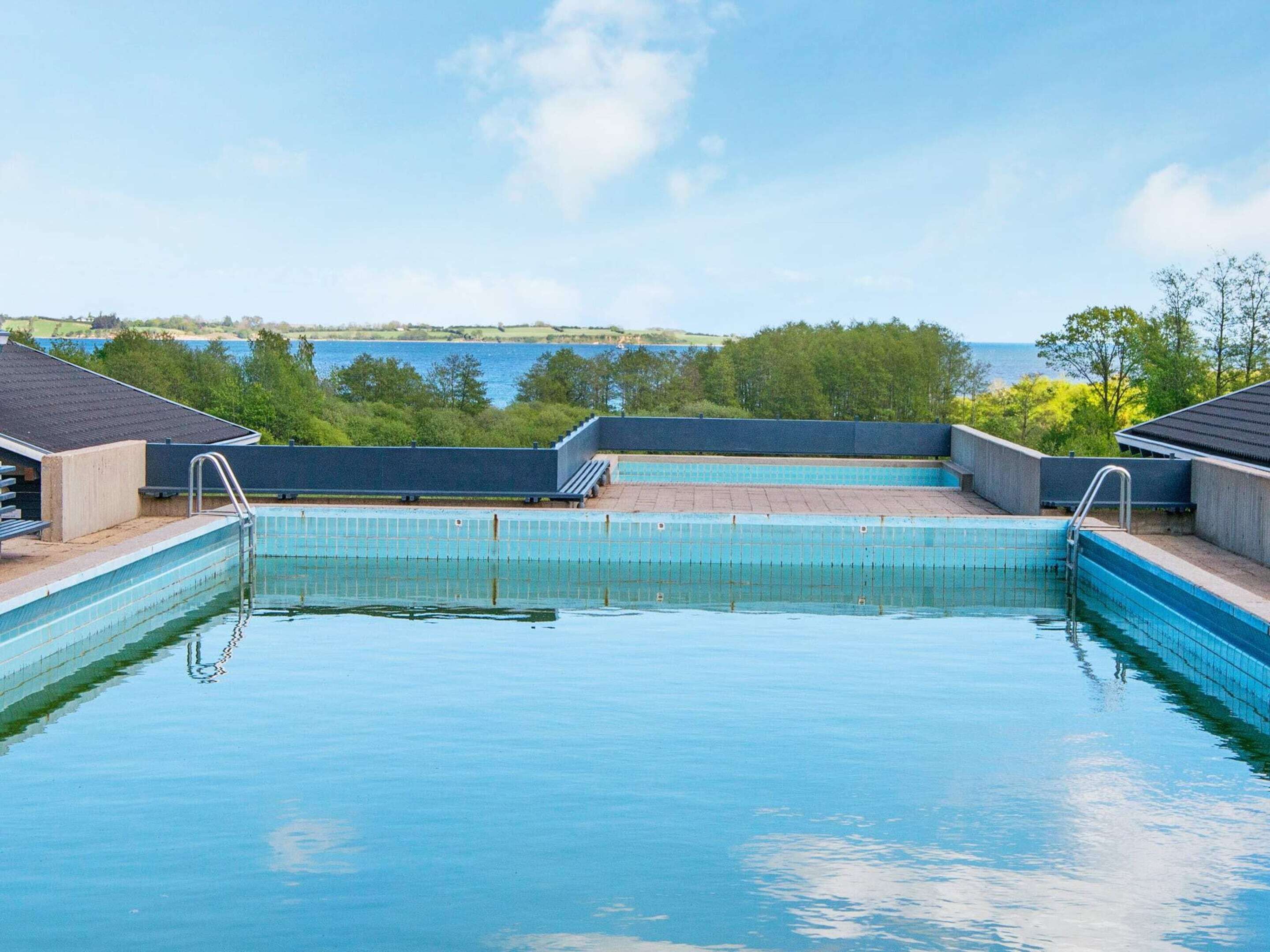 Uma piscina com água azul cristalina, rodeada por um deck de concreto, com vista para um corpo d'água e árvores ao fundo.