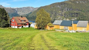 Une maison rouge avec un toit en pente et un petit porche, entourée de champs verdoyants et avec des montagnes en arrière-plan.