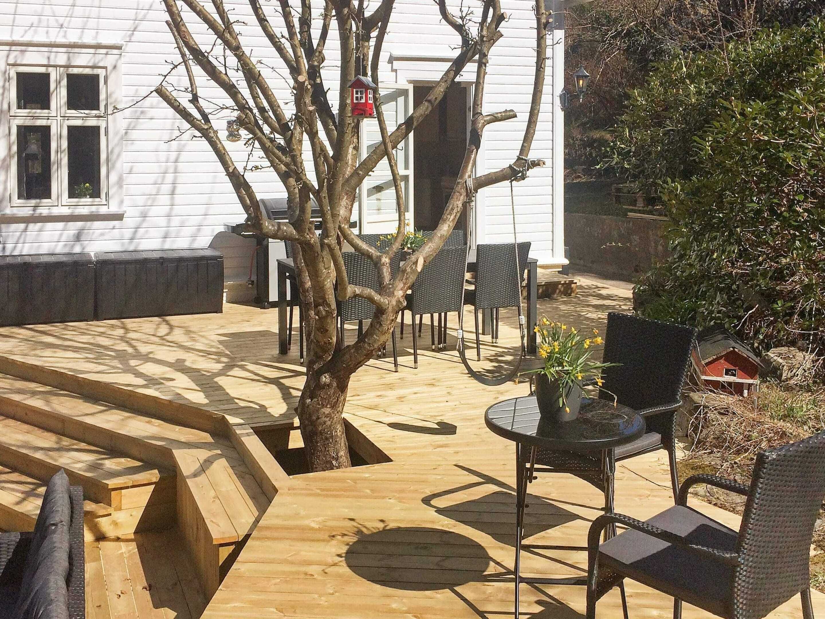 A wooden deck with a tree, outdoor furniture, and a white building in the background.