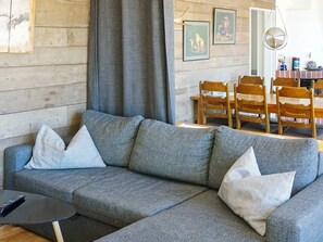 A grey sofa with pillows, a small black table, and a wooden dining table with chairs in a room with wooden walls and a hanging light fixture. - 8 Person Holiday Home in Hyltebruk (Hyltebruk)