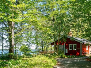A red cabin with a porch, surrounded by lush greenery and a body of water in the background. - 8 Person Holiday Home in Hyltebruk (Hyltebruk)
