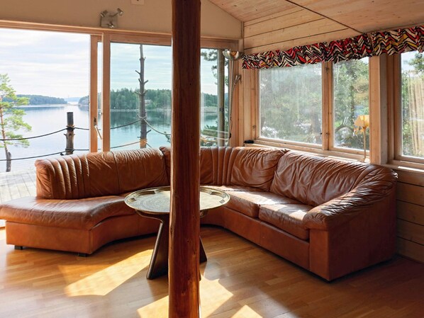 A room with a curved leather sofa, a round wooden table, and large windows offering a view of a lake and trees. - Holiday Home in Henån (Henån)