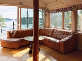 Una habitación acogedora con un sofá de cuero curvo, una mesita redonda y ventanas amplias con vistas a un lago y árboles.