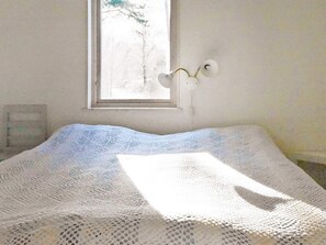 A room with a bed covered by a white knitted blanket, a window with a view of trees, and a wall-mounted light fixture. - Holiday Home in Henån (Henån)