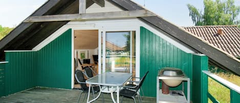 A small, green-roofed building with a sloped roof, a glass door entrance, a table with chairs, and a grill outside.
