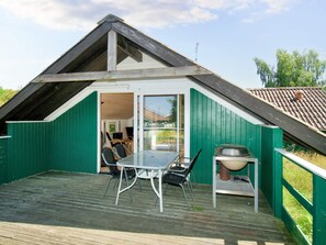 A small, green-roofed building with a sloped roof, a glass door entrance, a table with chairs, and a grill outside. - 6 Person Holiday Home in Skibby (Skibby)