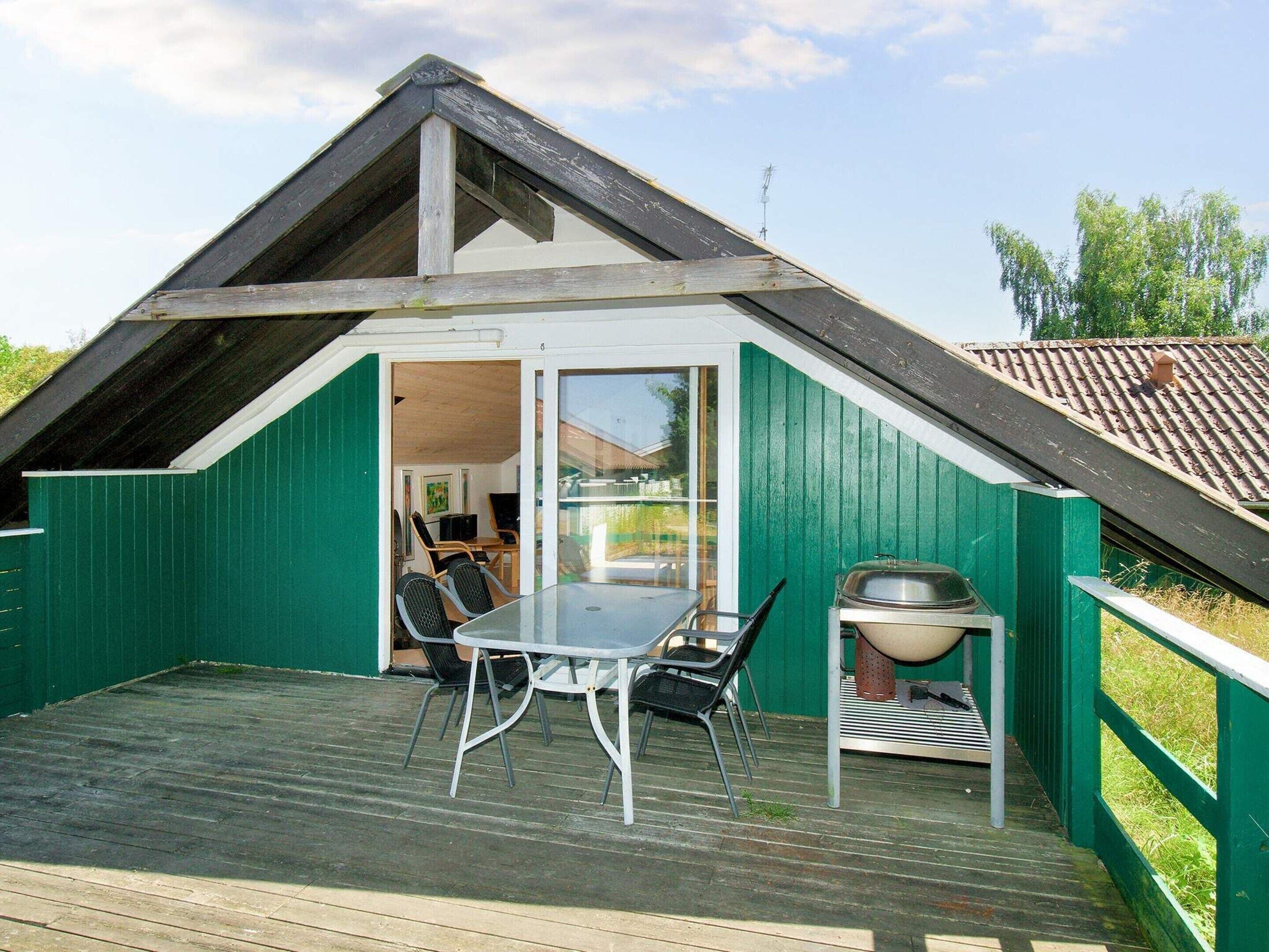 A small, green-roofed building with a sloped roof, a glass door leading inside, a table with chairs, and a grill outside.