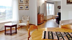 A living room with a wooden dining table, a floral armchair, and a sofa. There are framed pictures on the wall and a view of the outside through a window. - 8 Person Holiday Home in Skiftun (Hjelmeland)