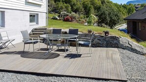 A wooden deck with outdoor furniture, a stone wall, and a garden with various plants and a view of a forested hill. - 8 Person Holiday Home in Skiftun (Hjelmeland)