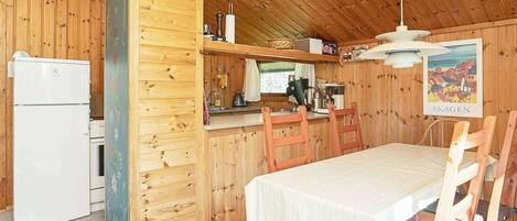 A compact kitchen with wooden walls, a white refrigerator, a dining table with chairs, and a hanging light fixture.