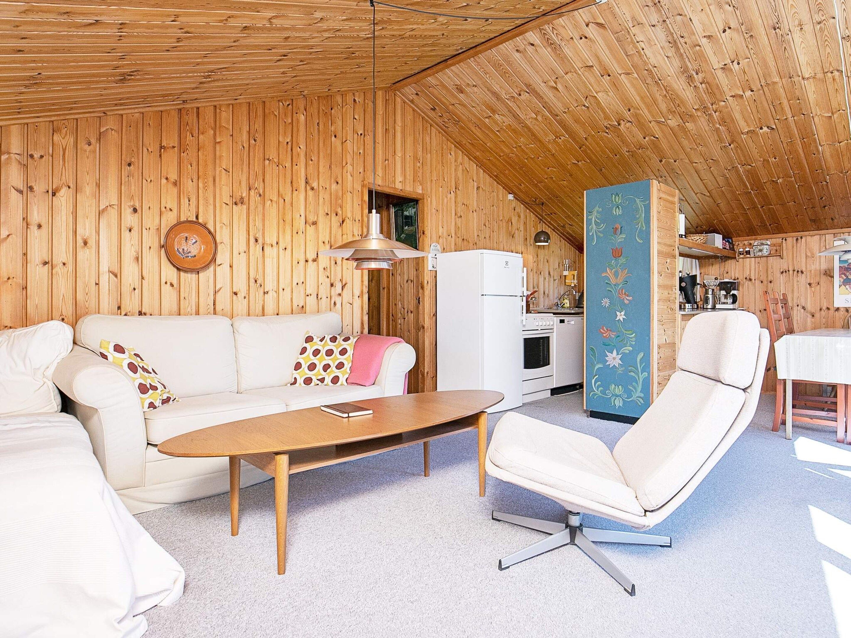 Ein gemütliches Zimmer mit Holzbalkendecke, einem weißen Sofa, einem Holztisch, einem weißen Sessel und einer Küchenzeile mit weißem Kühlschrank und Herd.