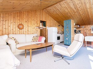 Une pièce chaleureuse avec un plafond en bois, un canapé blanc, une table basse en bois, un fauteuil blanc et un coin cuisine avec un réfrigérateur et une cuisinière blancs.
