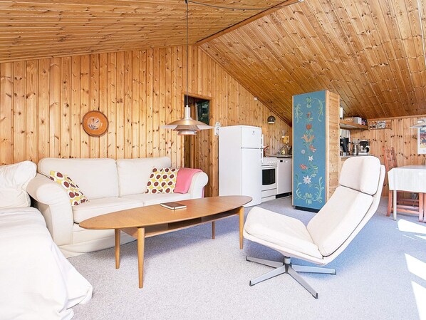 A cozy room with a wooden ceiling, a white sofa, a wooden coffee table, a white armchair, and a kitchen area with a white refrigerator and stove.