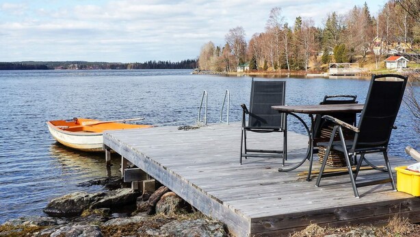 A wooden dock extending into a lake with a small boat, a table with chairs, and a view of a forested shoreline. - 4 Person Holiday Home in Ormaryd (Ormaryd)