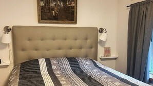 Een slaapkamer met een gestoffeerd hoofdeinde, een bed met een patroon, een lijst met een schilderij boven het bed, bedlampen en een raam met gordijnen.