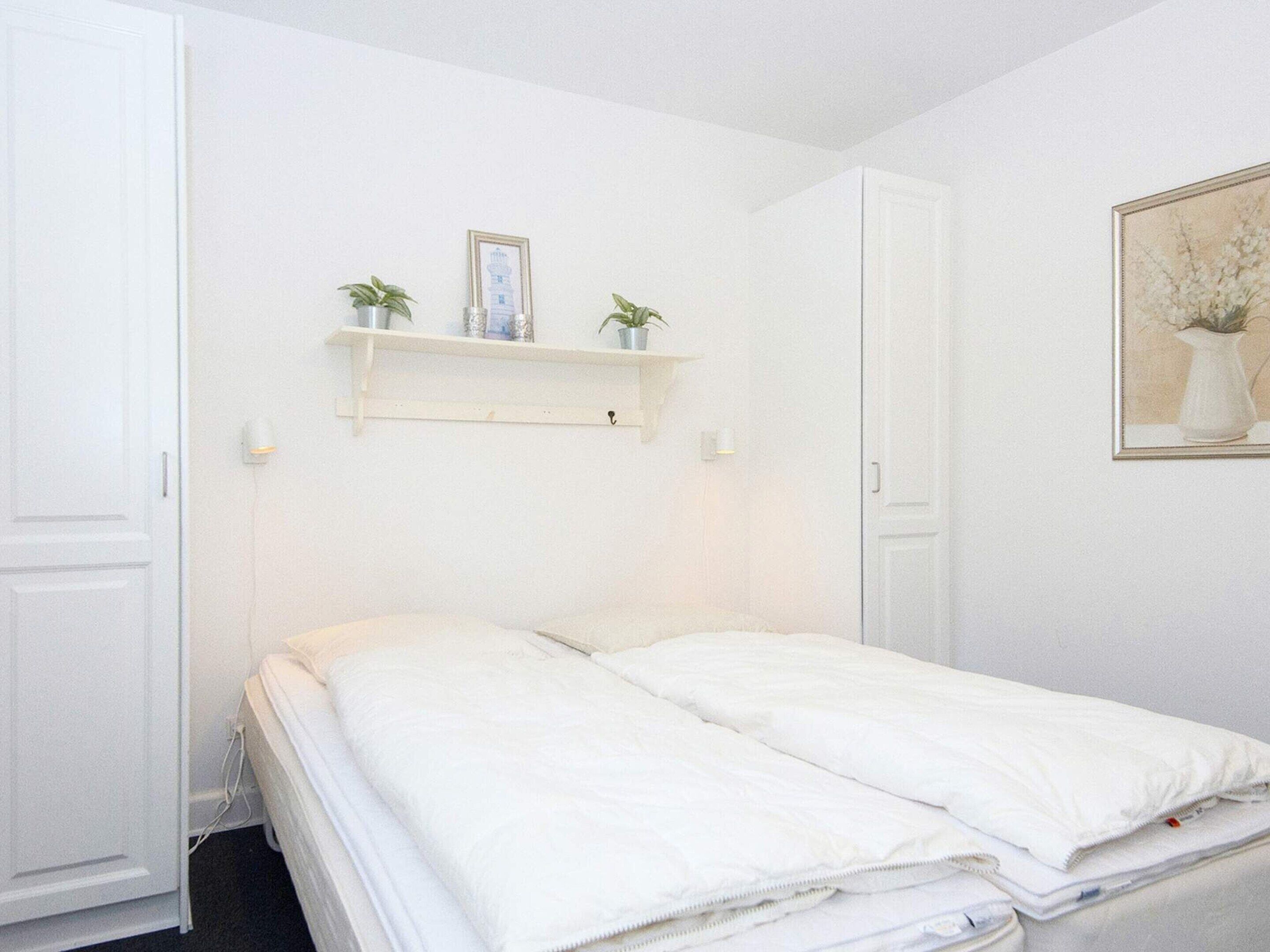 A bedroom with a bed, two white doors, a white wall, a shelf with two potted plants, and a framed picture.