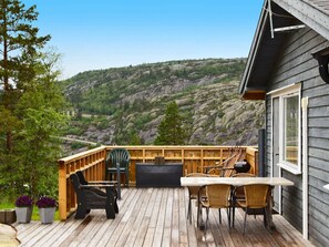 Une terrasse en bois avec des meubles d’extérieur, une grande fenêtre et une vue sur une colline rocheuse.