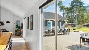 Interior moderno com uma longa mesa de jantar de madeira, um espaço de estar acolhedor e vista para uma casa de madeira e árvores.