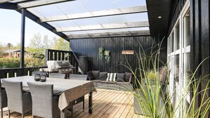An outdoor patio with a wooden deck, a grill, wicker furniture, and a glass enclosure. - 4 Star Holiday Home in Hals (Hals)