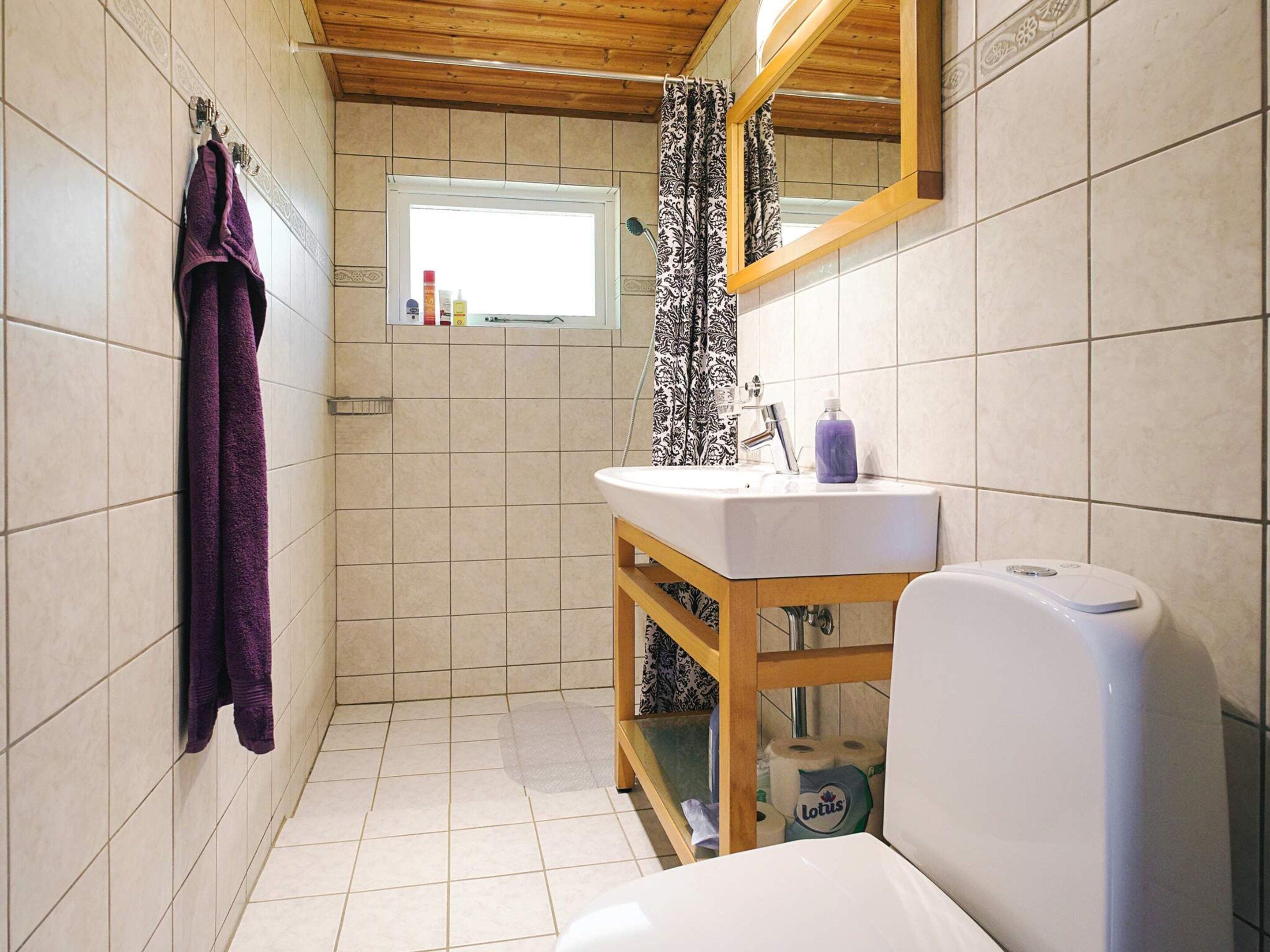 Baño con lavamanos blanco, espejo y un inodoro. Hay un perchero con una toalla morada en la pared.