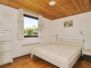 A bedroom with a wooden ceiling, a bed, a bedside table, a lamp, a window with curtains, and a radiator.