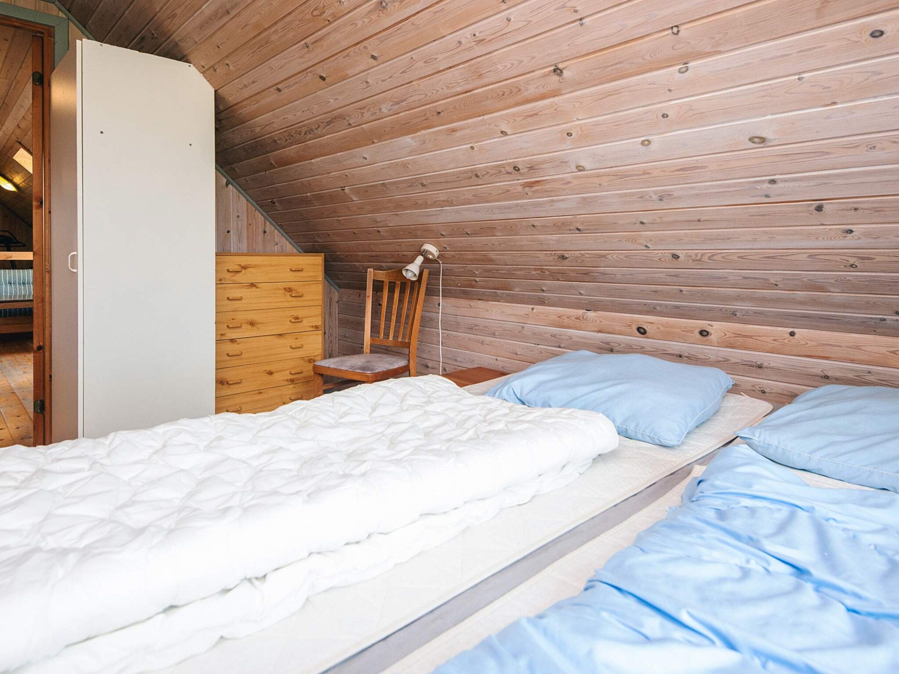 A bedroom with a wooden sloped ceiling, a single bed with white bedding, a wooden dresser, and a chair with a lamp.