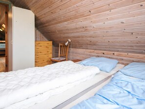 A bedroom with a wooden sloped ceiling, a single bed with white bedding, a wooden dresser, and a chair with a lamp.