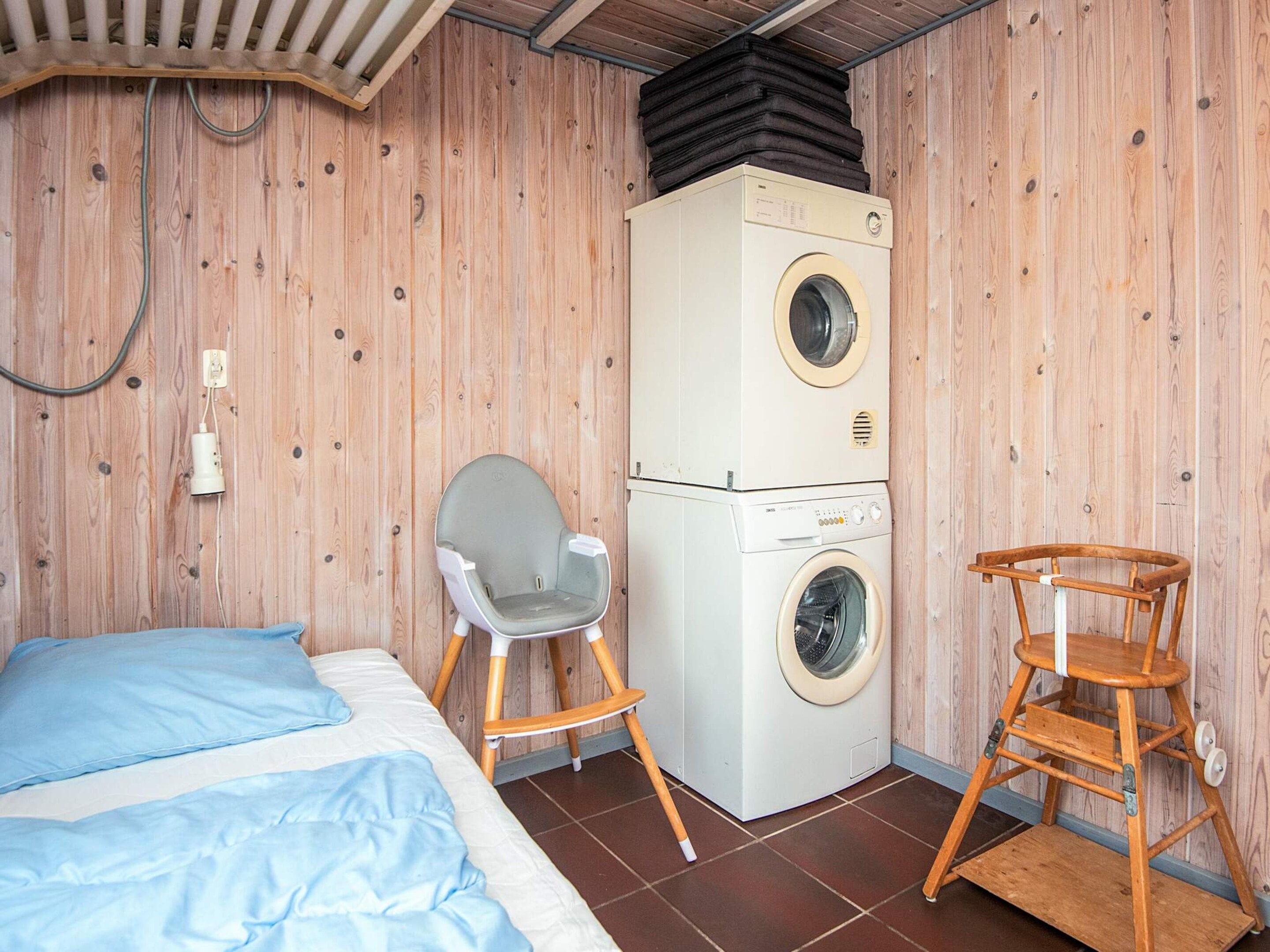 A compact room with a bed, a washing machine, a chair, and a wooden stool.