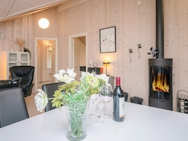 Una habitación con una mesa blanca, un jarrón con flores, copas de vino y una botella de vino. Hay una chimenea encendida y un reloj en la pared.