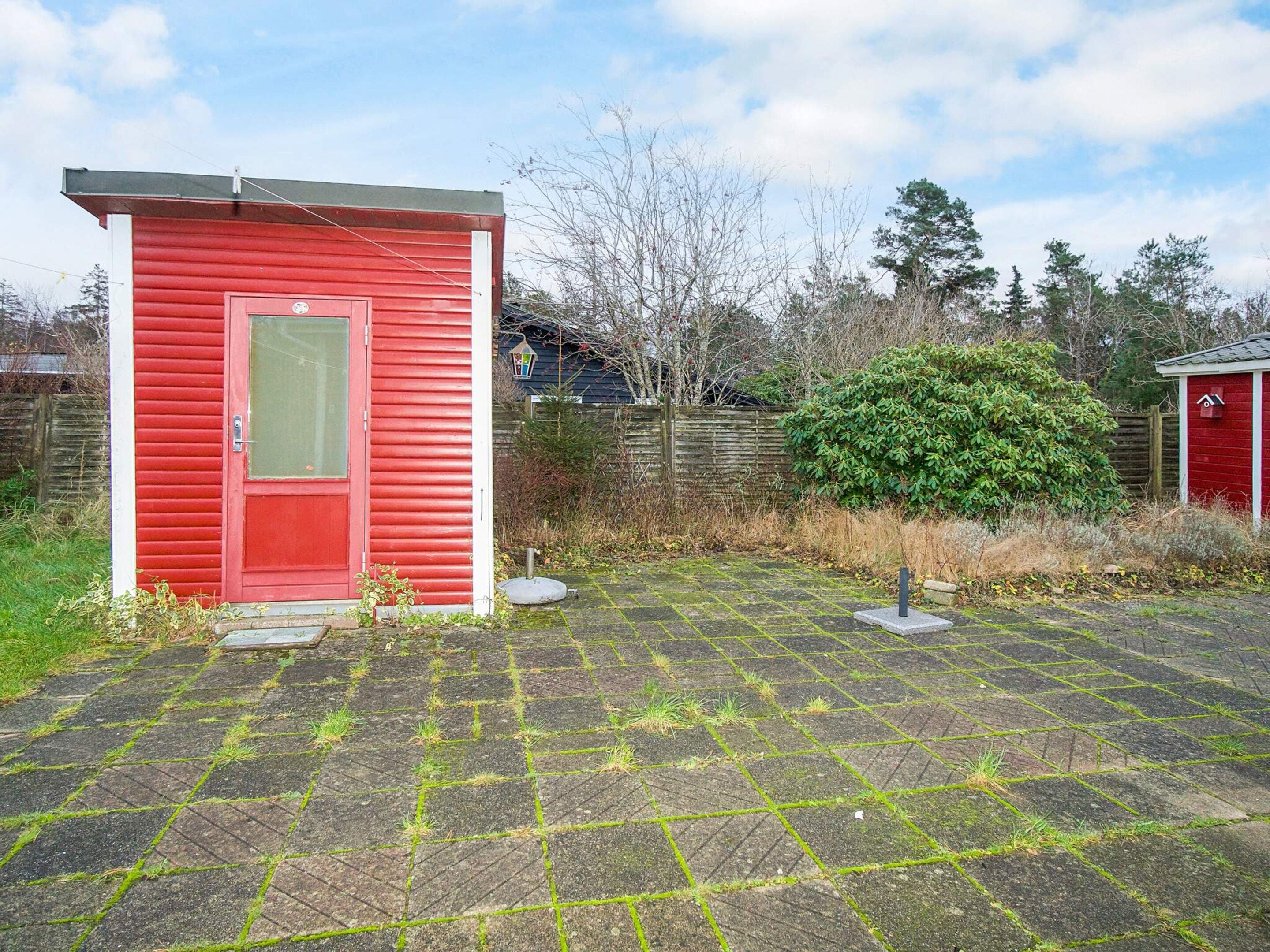 A red shed with a door and window, a paved area with grass patches, and a wooden fence in the background.
