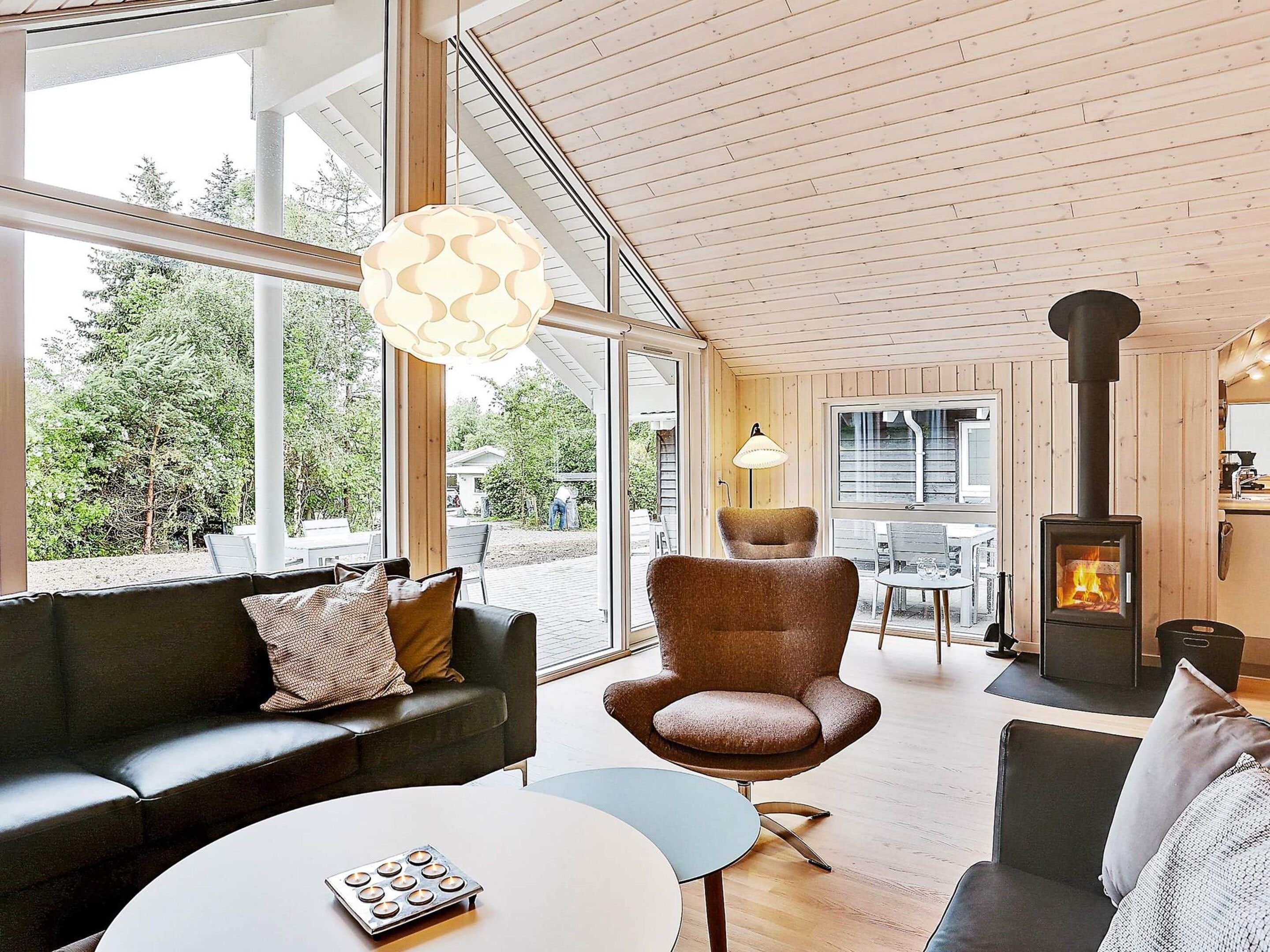 A modern interior with a sofa, armchair, and round table, a fireplace, and large windows offering a view of the outdoors.