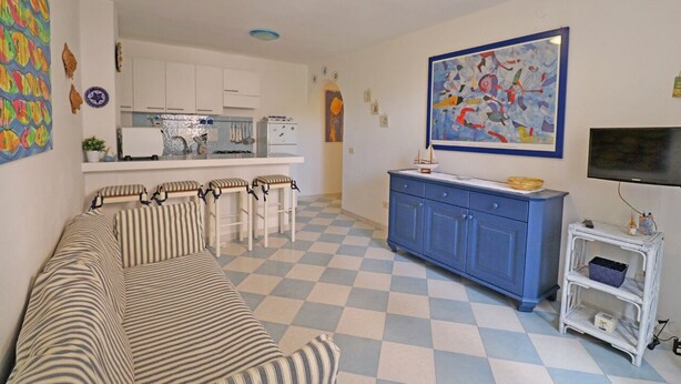 Un salon avec un canapé à rayures, un coin repas avec une cuisine en arrière-plan, un meuble bleu et un tableau au mur.