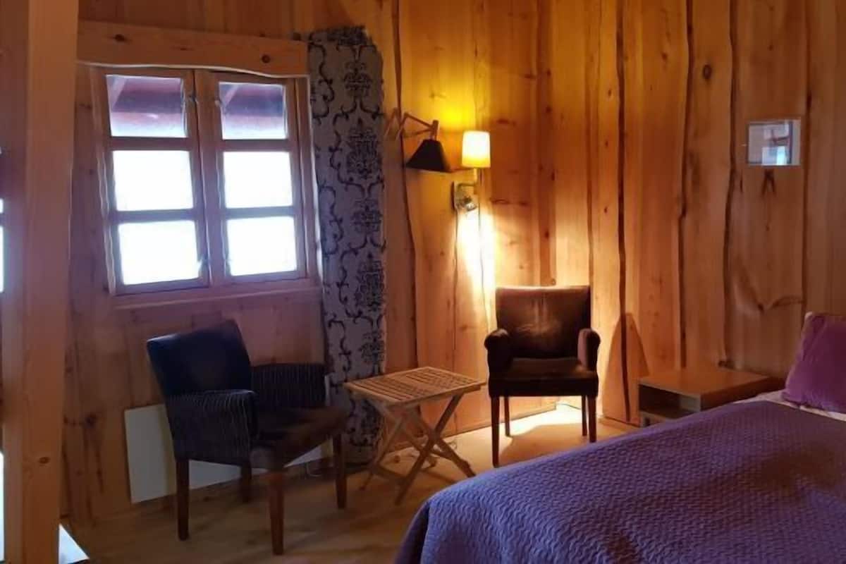 A wooden cabin room with a bed, two chairs, a small table, and a window.