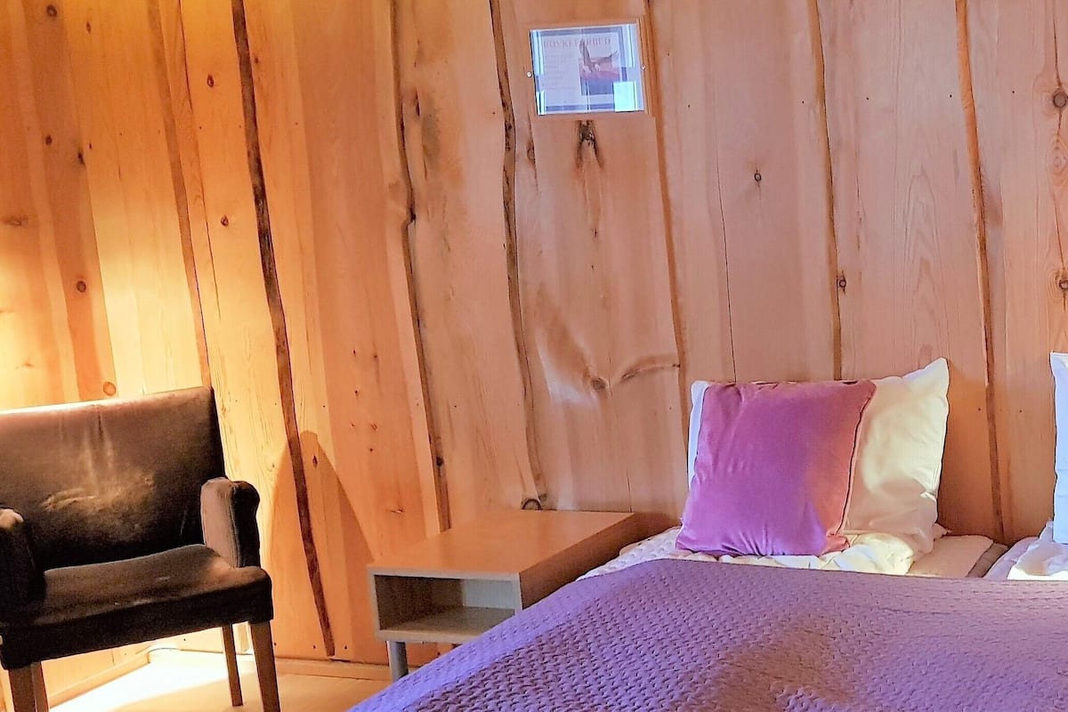 A wooden room with a bed, pillows, a chair, and a small table.