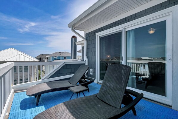 A balcony with two lounge chairs and a table, a sliding glass door, and a view of neighboring houses.