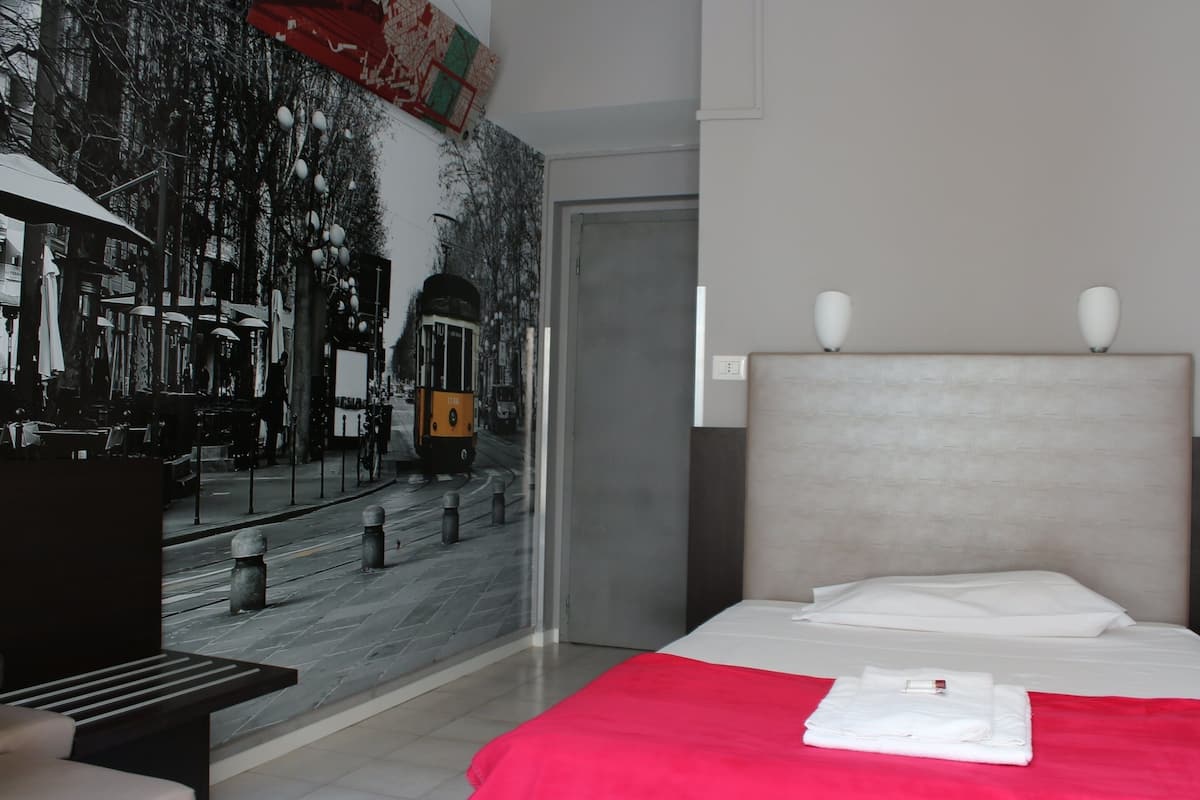 A hotel room with a bed, a red blanket, a neatly folded white towel, and a wall mural of a tram.