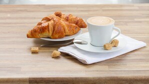 Una mesa de madera con un croissant, una taza de café y una cuchara.