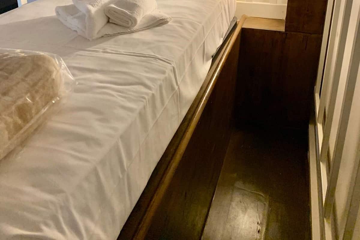 A single bed with white linens, a folded towel, and a plastic-wrapped item on the bed.