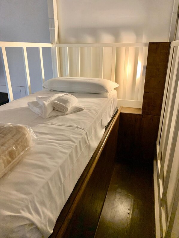 Una cama individual con sábanas blancas, una toalla doblada y un objeto envuelto en plástico sobre la cama.