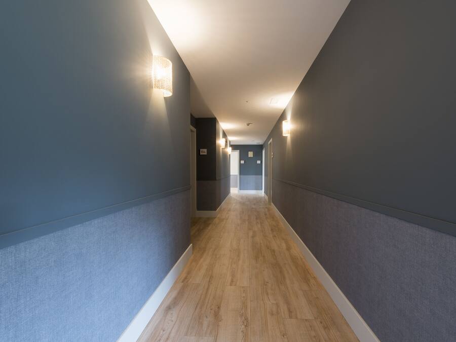 Un pasillo estrecho con suelo de madera y paredes grises, con luces empotradas en la pared y una puerta al final.