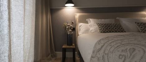 Un dormitorio ordenado con cama, mesita de noche, lámpara y un florero con flores secas.