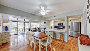 Una cocina amplia con una isla central, un comedor con mesa de madera y sillas blancas, y un amplio ventanal corredero que da a un balcón.
