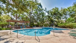 Una piscina con sillones de descanso y un pabellón, rodeada de árboles.