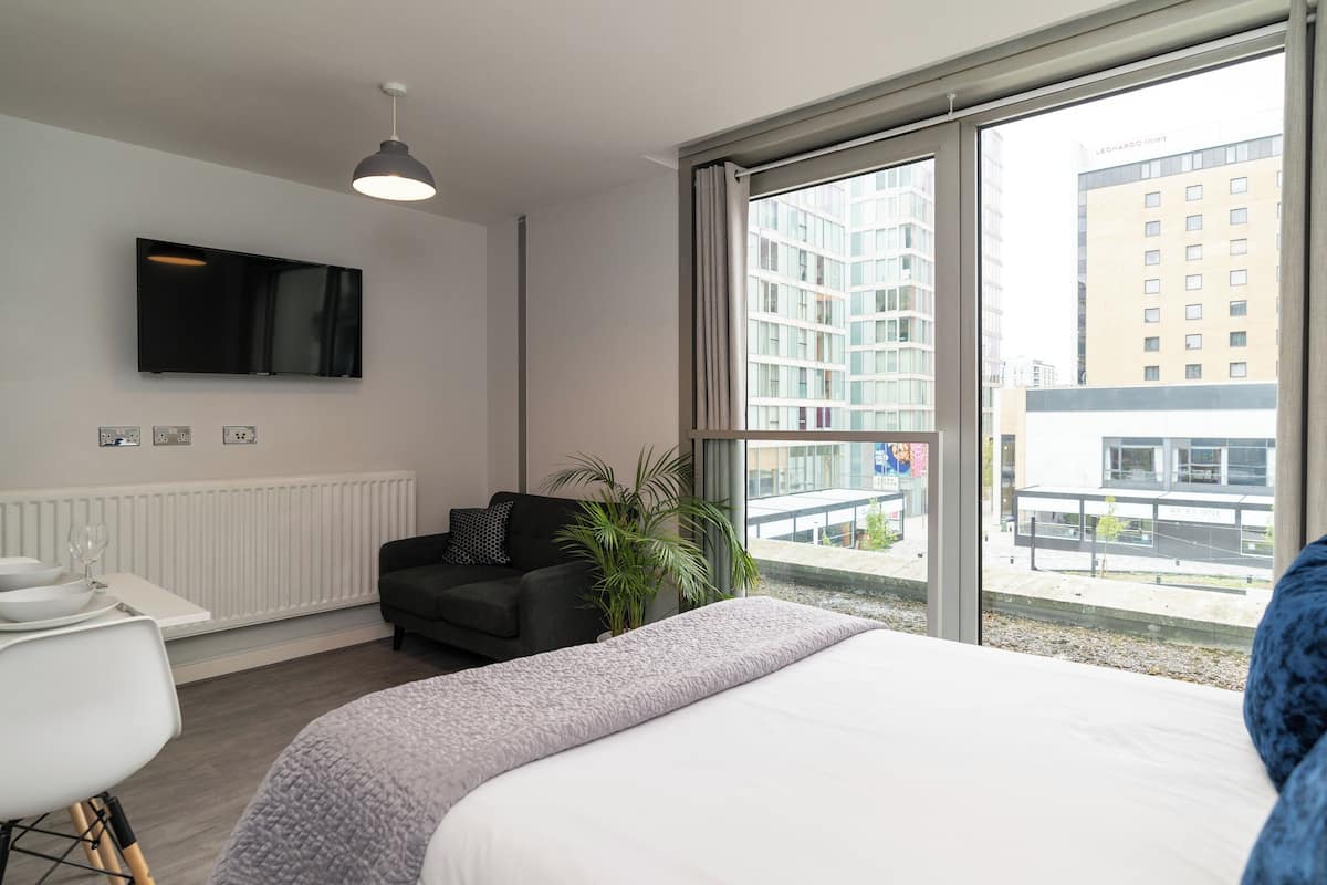 A modern hotel room with a large bed, a desk with a chair, a television mounted on the wall, and a large window offering a city view.