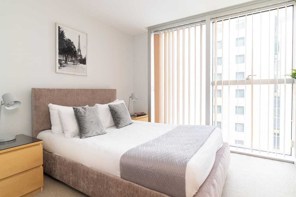 A modern bedroom with a large bed, bedside tables, a nightstand, a lamp, a framed picture of the Eiffel Tower, and a view of a building through floor-to-ceiling windows.