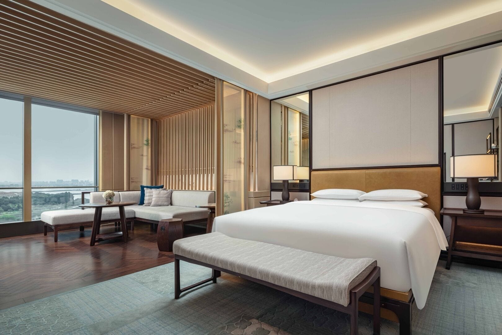 A modern hotel room with a large bed, a seating area with a bench, a sofa, and a view of the cityscape.