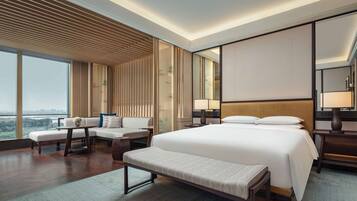 A modern hotel room with a large bed, a seating area with a bench, a sofa, and a view of the cityscape.
