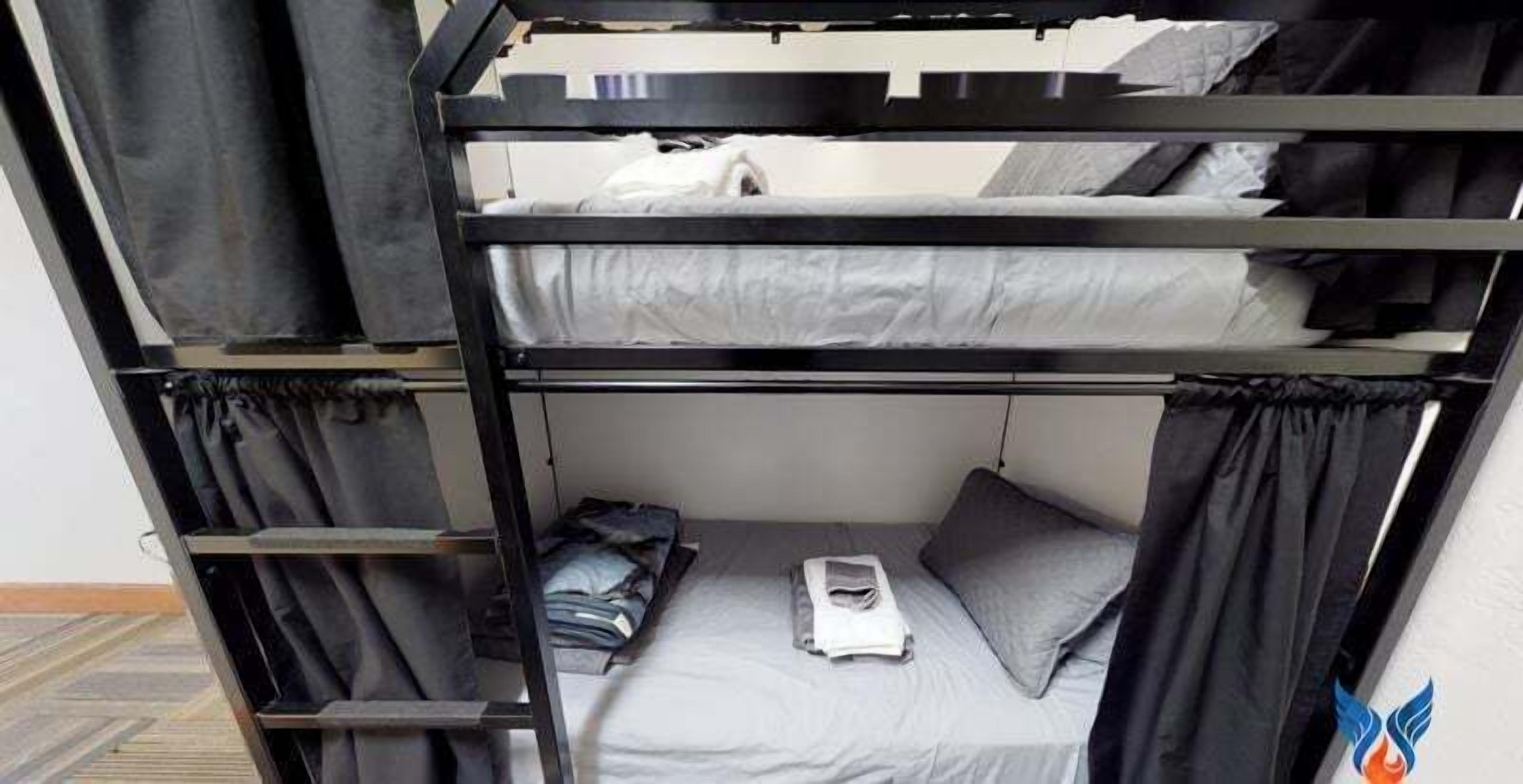 Ein Etagenbett mit einem unterem und einem oberen Bett, beide mit grauer Bettwäsche. Auf dem unteren Bett liegen ein gefaltetes Handtuch und ein Buch.